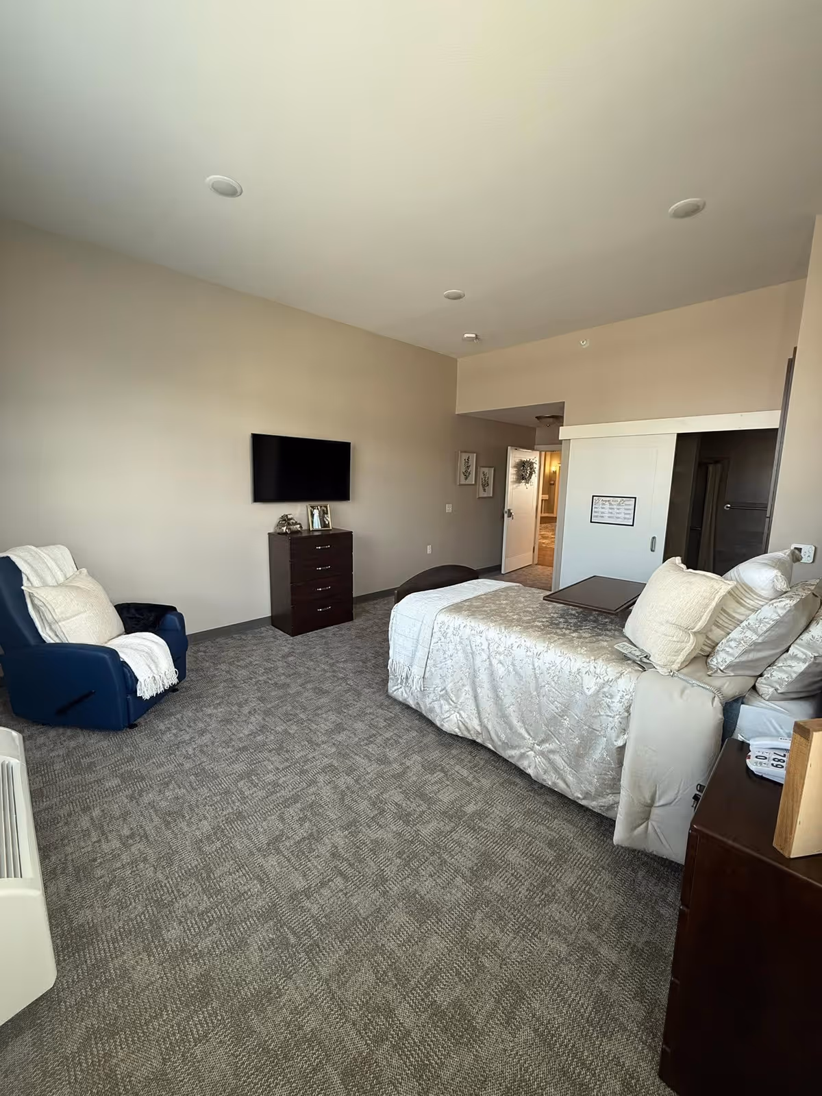 Well-lit senior living bedroom with a bed, dresser, wall-mounted TV, and blue chair.