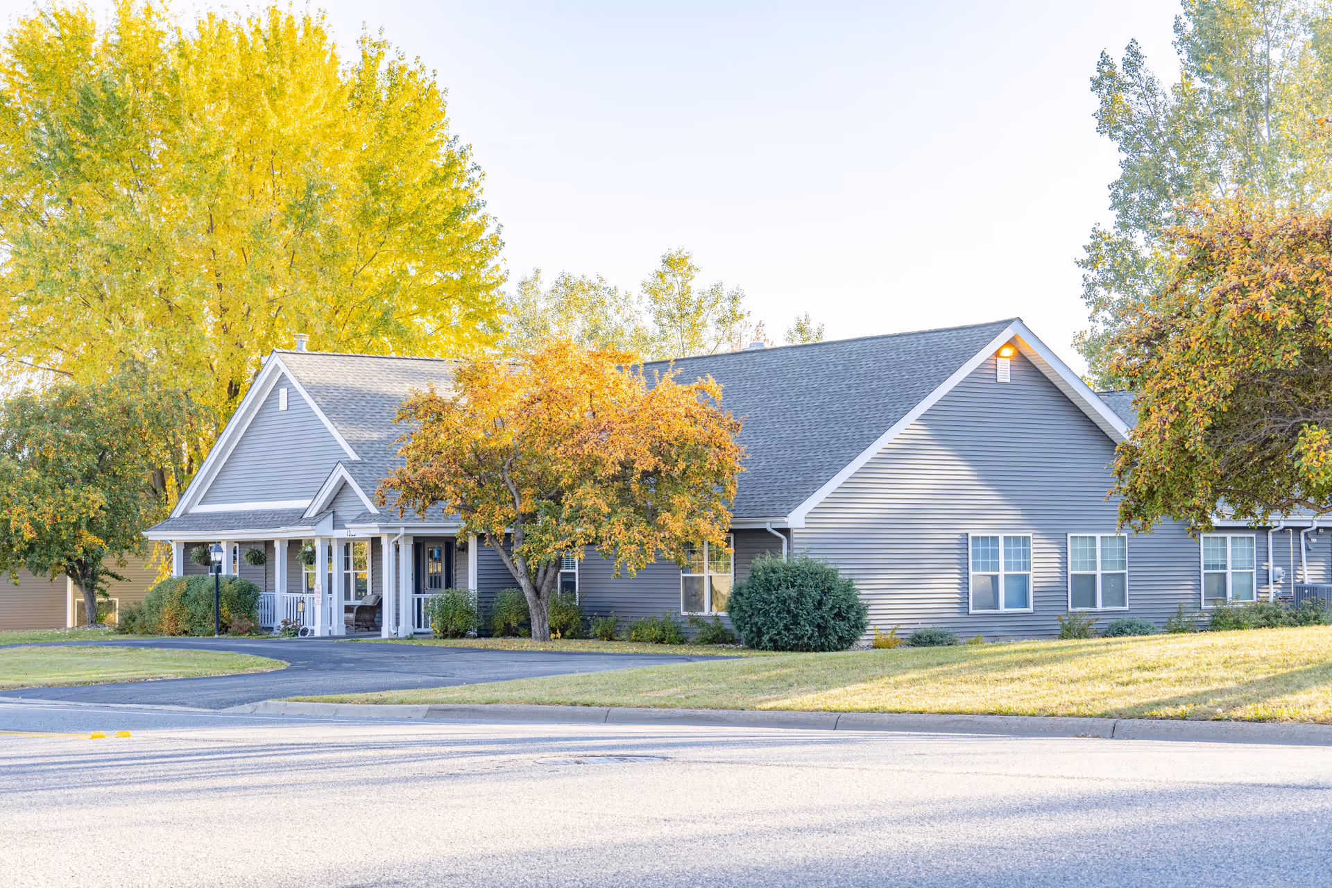 Exterior view of a single-story gray building with a gabled roof, surrounded by trees with autumn foliage and a well-maintained lawn, under a clear sky.