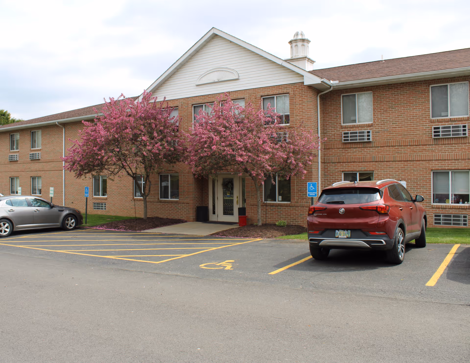 Exterior view of a two-story brick building with a peaked roof and white trim. Two pink flowering trees flank the entrance, which has glass double doors. There are several windows with air conditioning units, a parking lot with two cars, and marked handicapped parking spaces in front of the building.