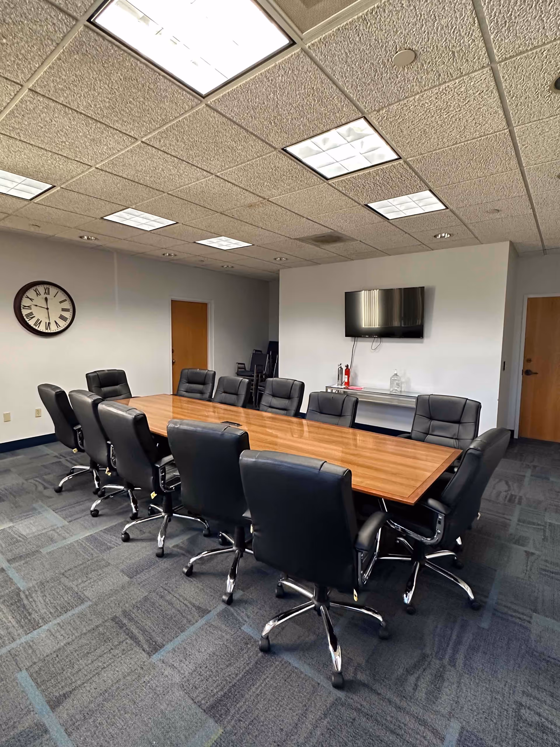 A conference room with a long wooden table, black leather chairs, and a wall-mounted TV.