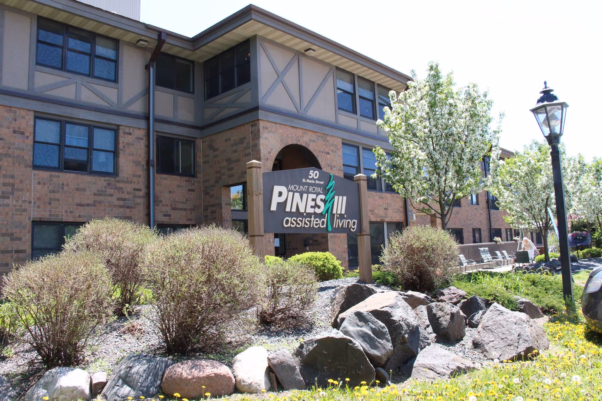 Brick exterior of Pines Assisted Living with landscaping, sign, and outdoor seating.