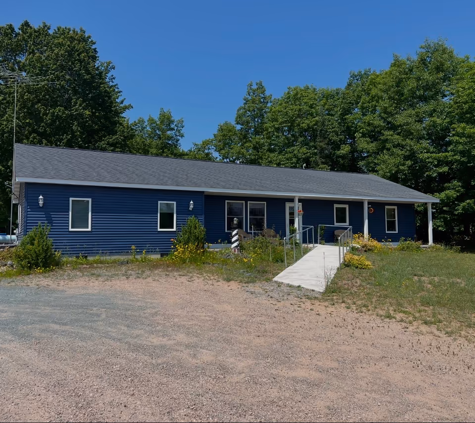 Blue single-story assisted living facility with ramp and surrounding trees.