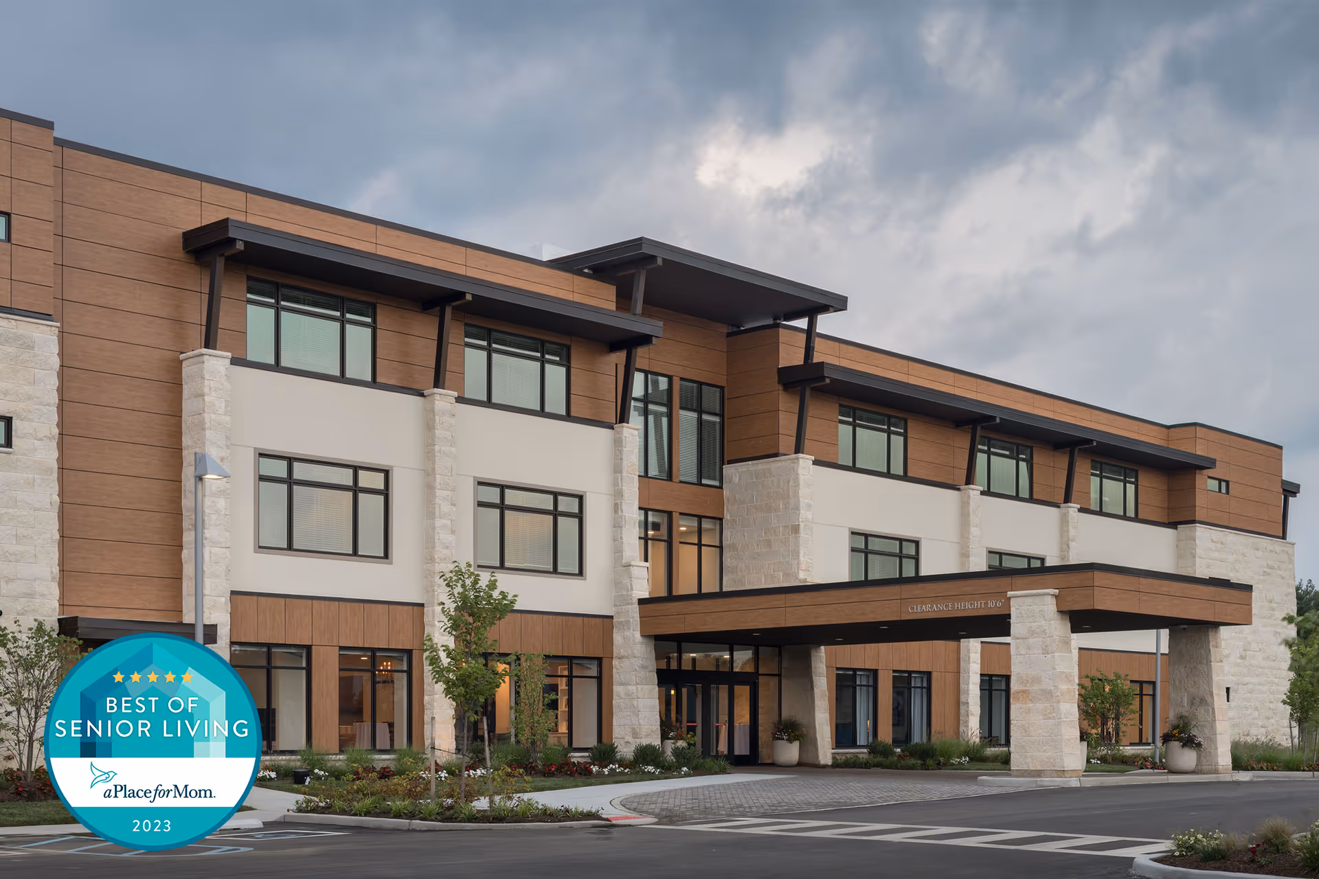 Modern three-story senior living building exterior with a covered entrance, landscaping, and a cloudy sky.