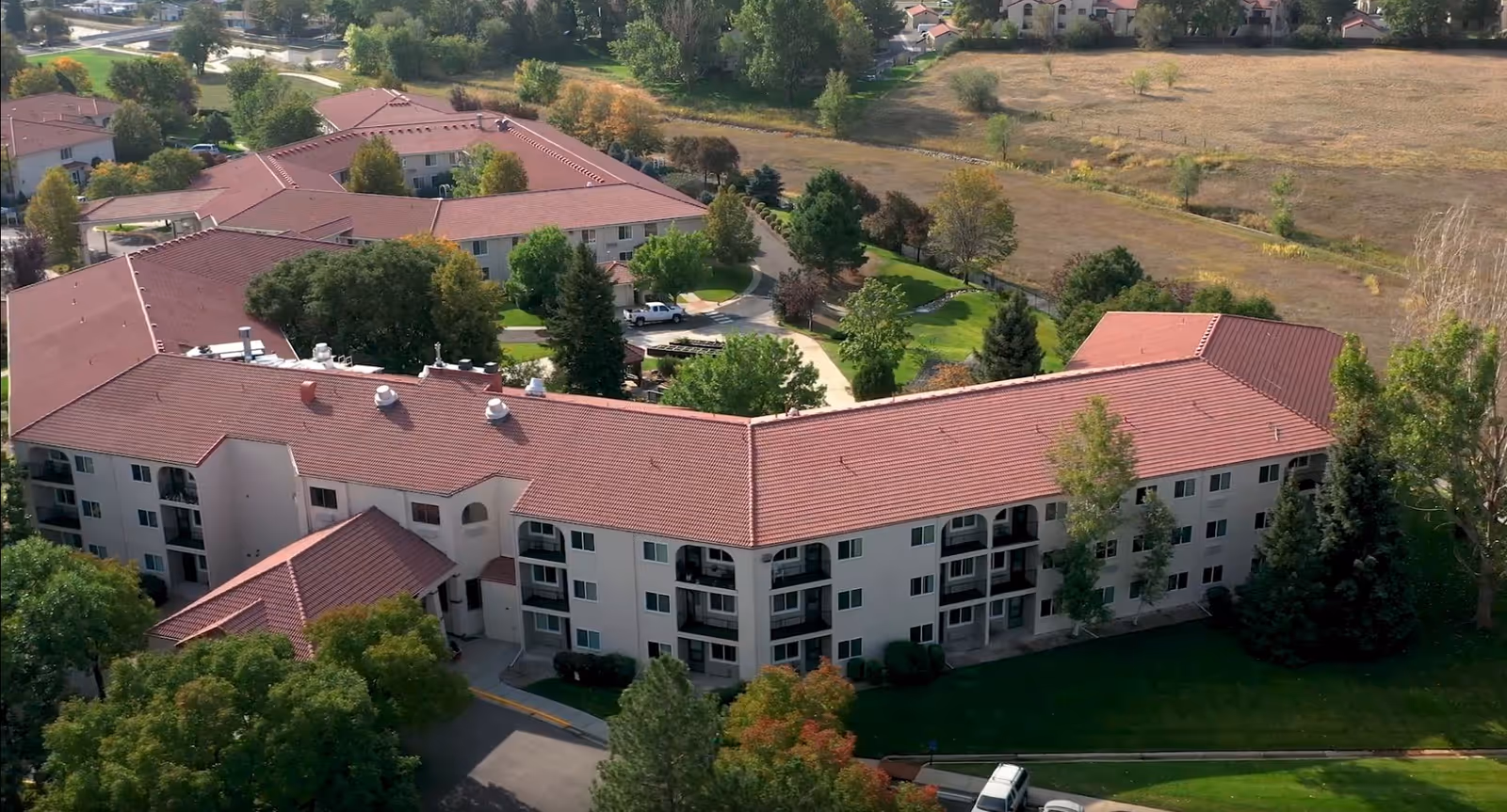 Aerial view of San Marino Retirement Community showing multiple connected buildings with red-tiled roofs surrounded by trees and green lawns, with a dry field in the background.