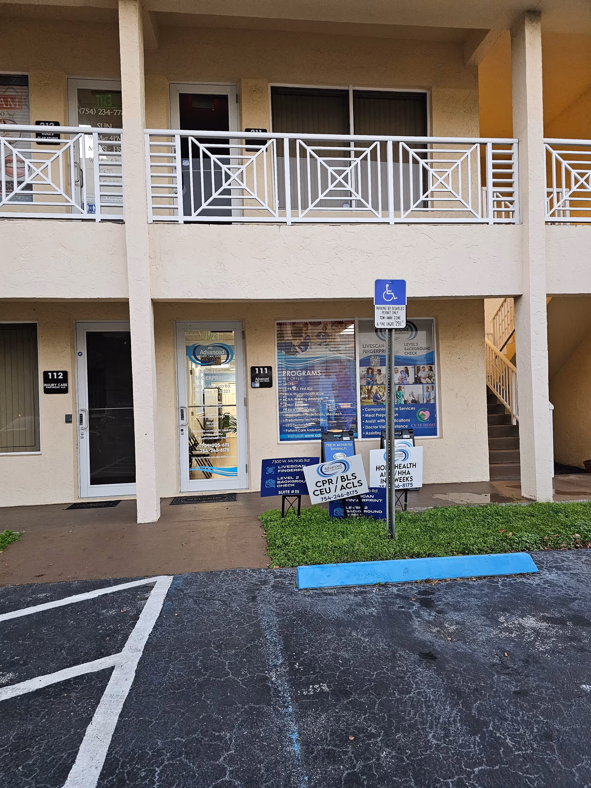 Two-story beige building exterior with entry doors, balcony railing, disabled parking sign, and promotional signs outside.