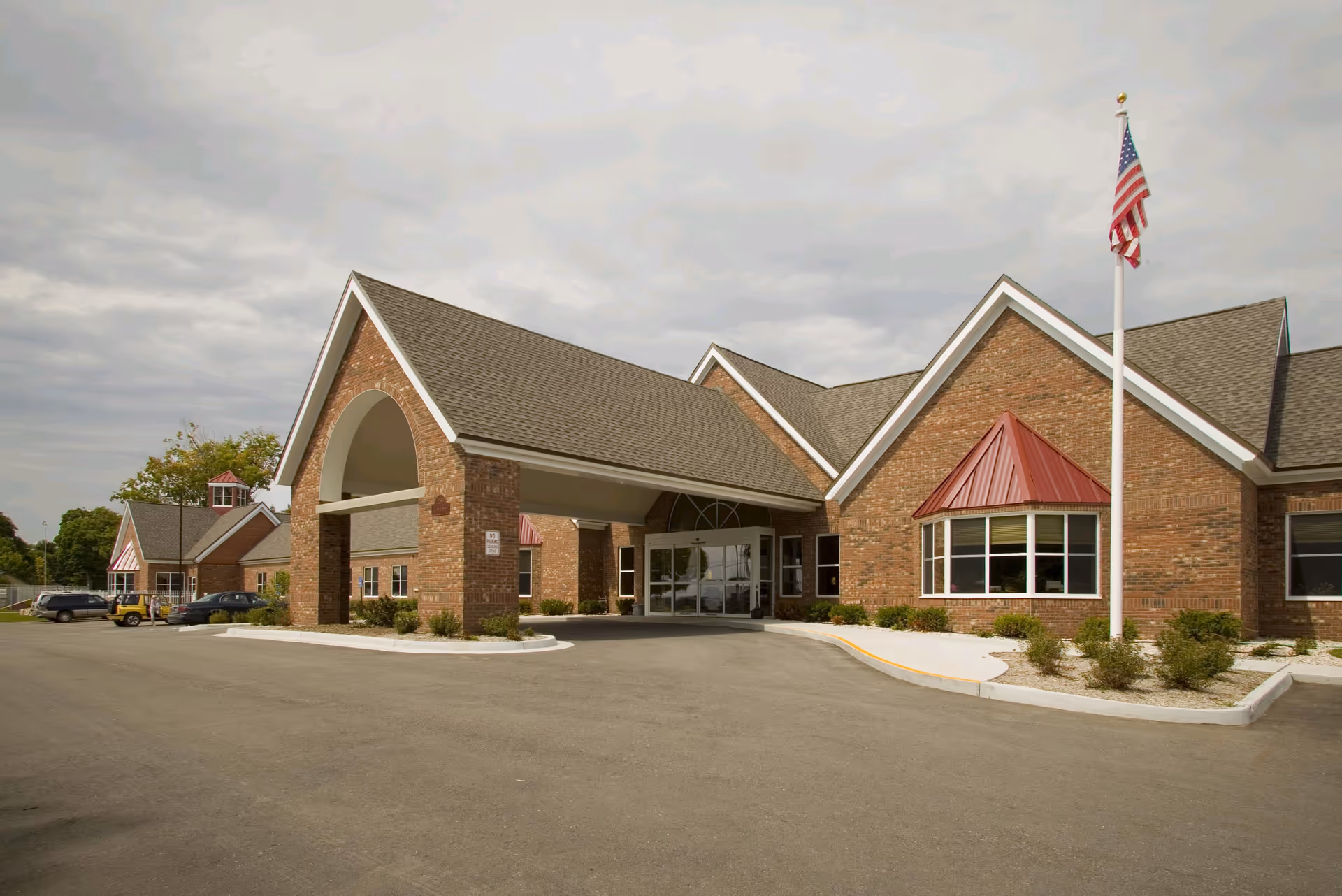 Brick senior living facility entrance with portico, flagpole, and surrounding parking lot.