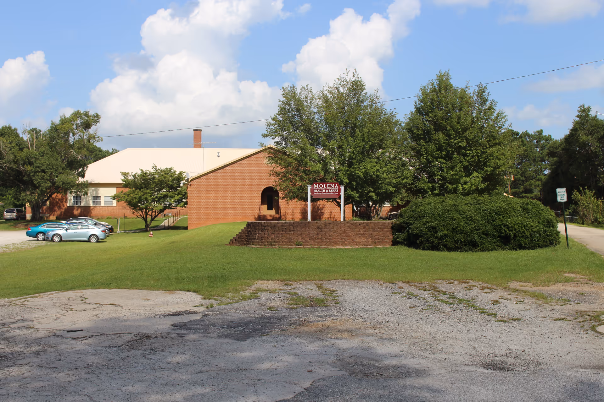 Brick Molena Health & Rehab building with sign, cars, and green lawn under blue sky.