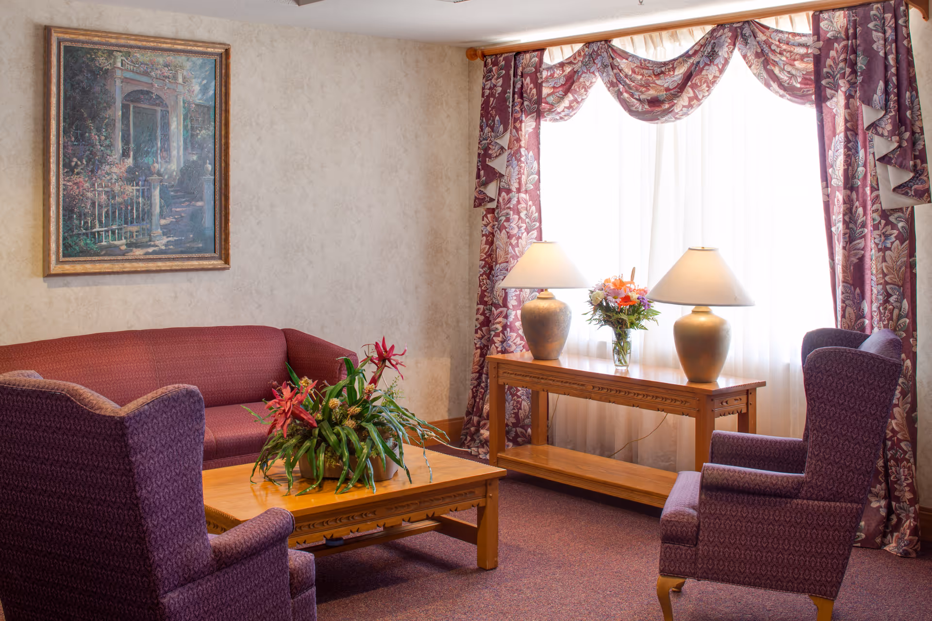 A cozy living room with a maroon sofa and two matching armchairs surrounding a wooden coffee table with a green plant centerpiece. Behind the coffee table is a wooden console table with two lamps and a vase of flowers. The window is adorned with sheer white curtains and purple floral drapes. A framed painting hangs on the wall above the sofa.
