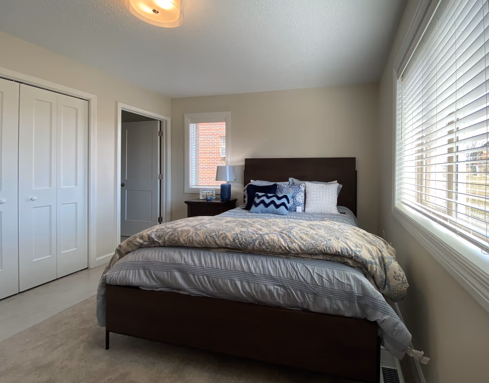 A bedroom with a neatly made bed featuring multiple pillows and a patterned comforter. There is a nightstand with a blue lamp and a small picture frame next to a window with blinds. The room has beige walls, a closet with white double doors, and a door leading to another room.