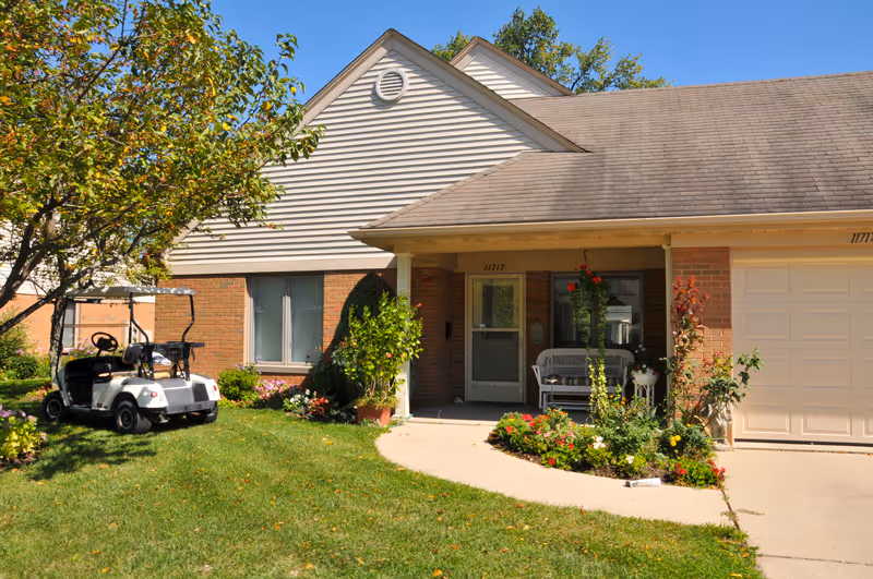 Exterior view of a single-story senior living community unit with a small porch, a white bench, and a garden with flowers. A golf cart is parked on the grass near the building. The building has brick and siding walls with a garage door on the right side.
