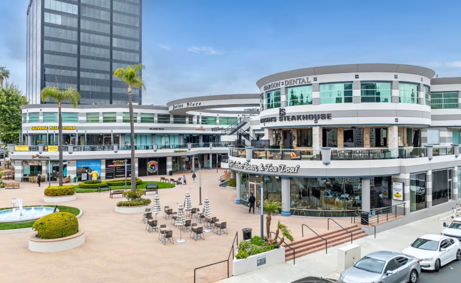 Exterior view of a modern shopping complex with outdoor seating and palm trees.