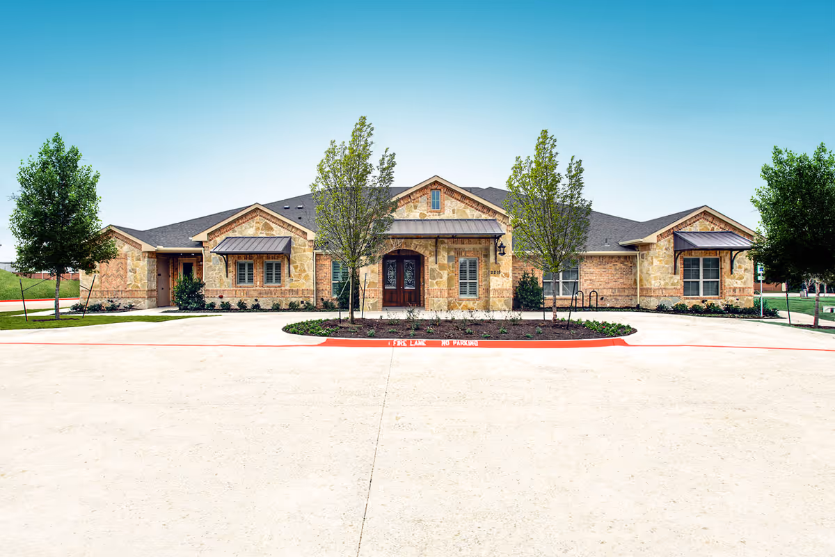 Front exterior view of a single-story building with stone and brick facade, multiple windows with awnings, a central entrance with double doors, and landscaped area with small trees and shrubs. The sky is clear and blue.
