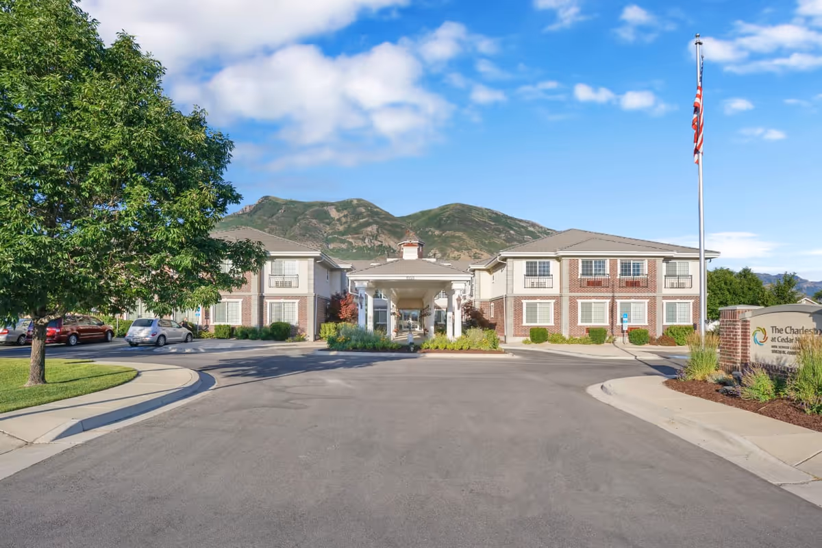Front exterior of The Charleston at Cedar Hills senior living building with a circular driveway, flagpole, landscaped entrance and mountains in the background.