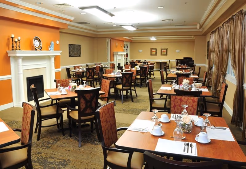 Dining room with multiple set tables and chairs, place settings, and a decorative fireplace in a senior living facility.
