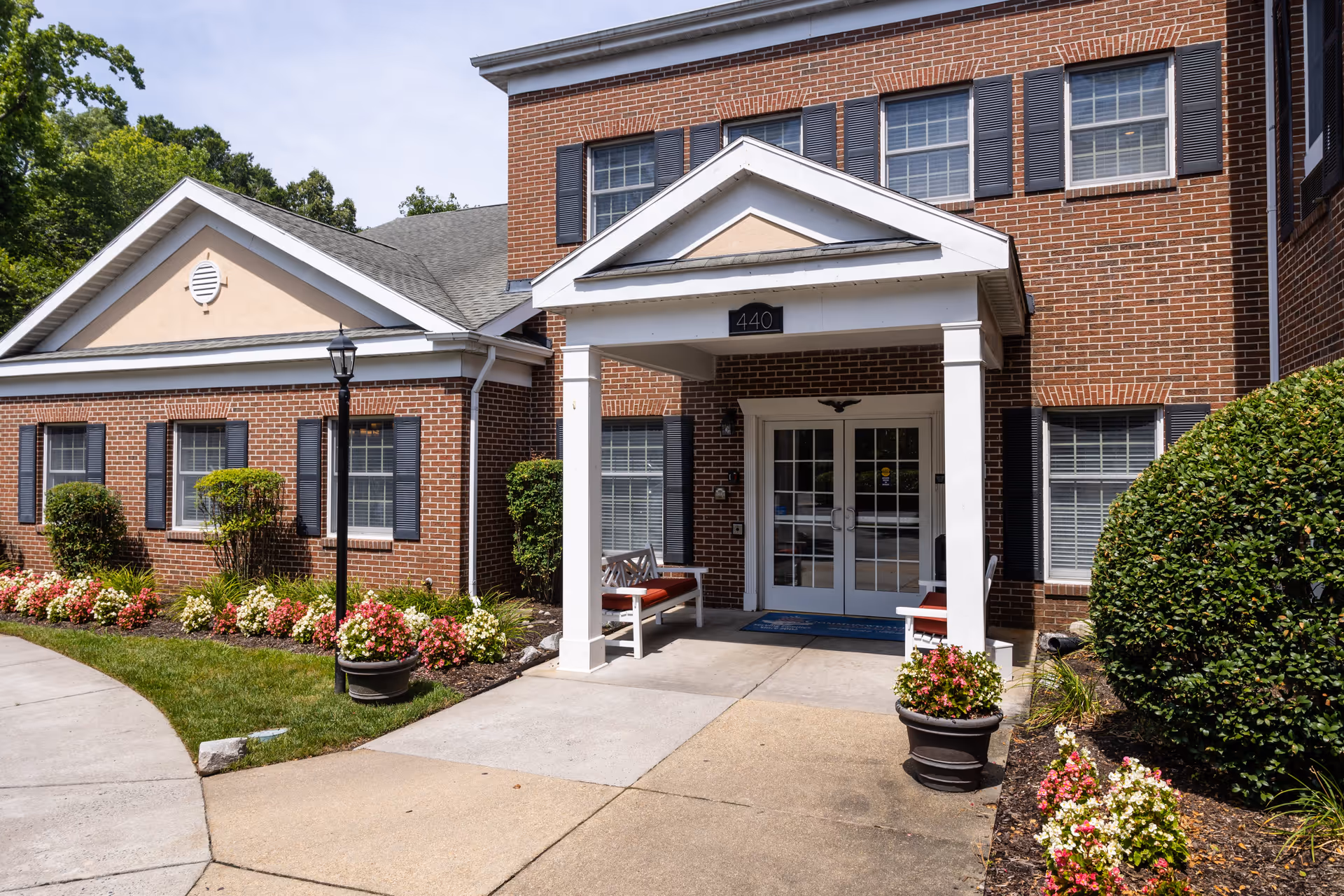 Entrance of a brick building with white columns and a small covered porch. There are two white benches with red cushions on either side of the entrance door. The building has multiple windows with black shutters. The walkway leading to the entrance is surrounded by well-maintained flower beds and green shrubs.