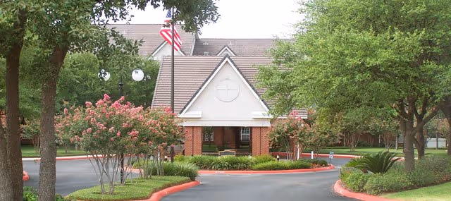 Circular driveway and landscaped entrance leading to the brick front of a residential building with flagpoles and trees.