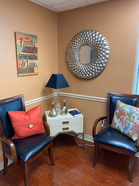 Two dark chairs with colorful pillows flank a white side table, lamp, and decorative wall art.