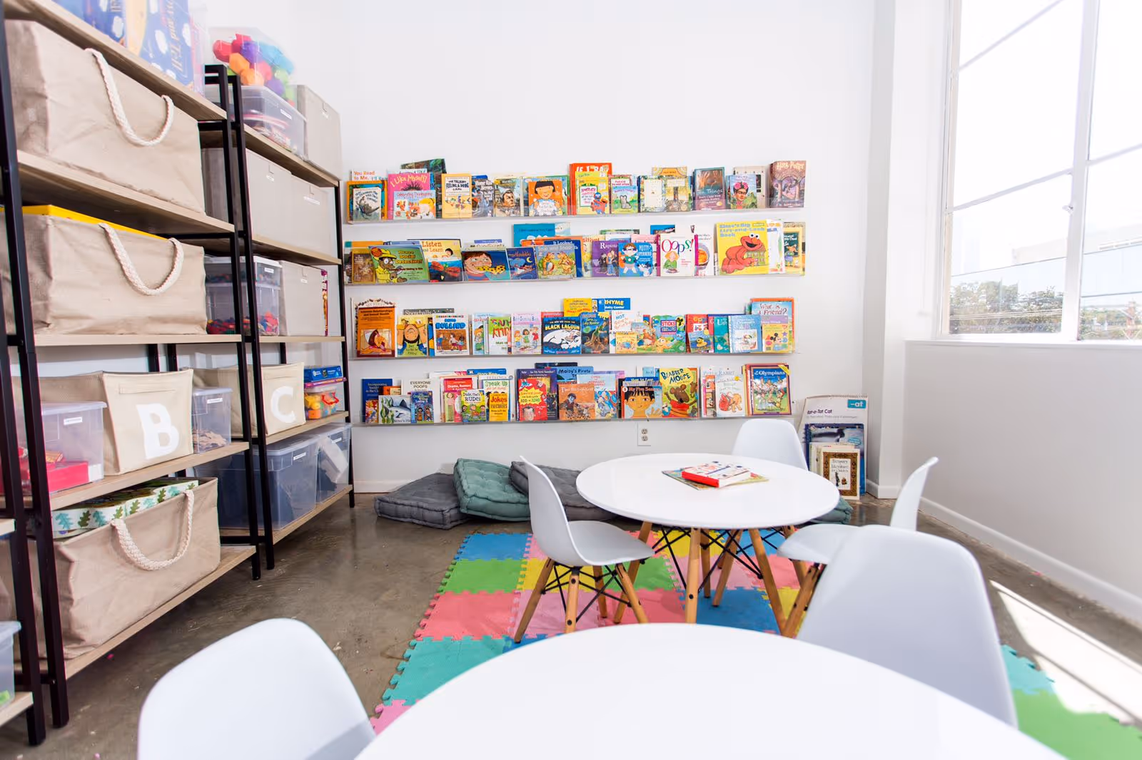 Bright children's reading area with bookshelves, colorful foam mats, and round tables.