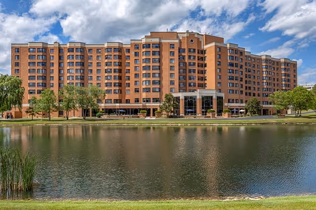 A large multi-story brick building with many windows, situated behind a pond with green grass and trees around it under a partly cloudy sky.