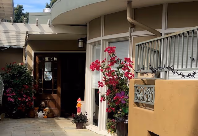 Entrance to a senior living facility with glass doors, potted flowers, and beige exterior walls.