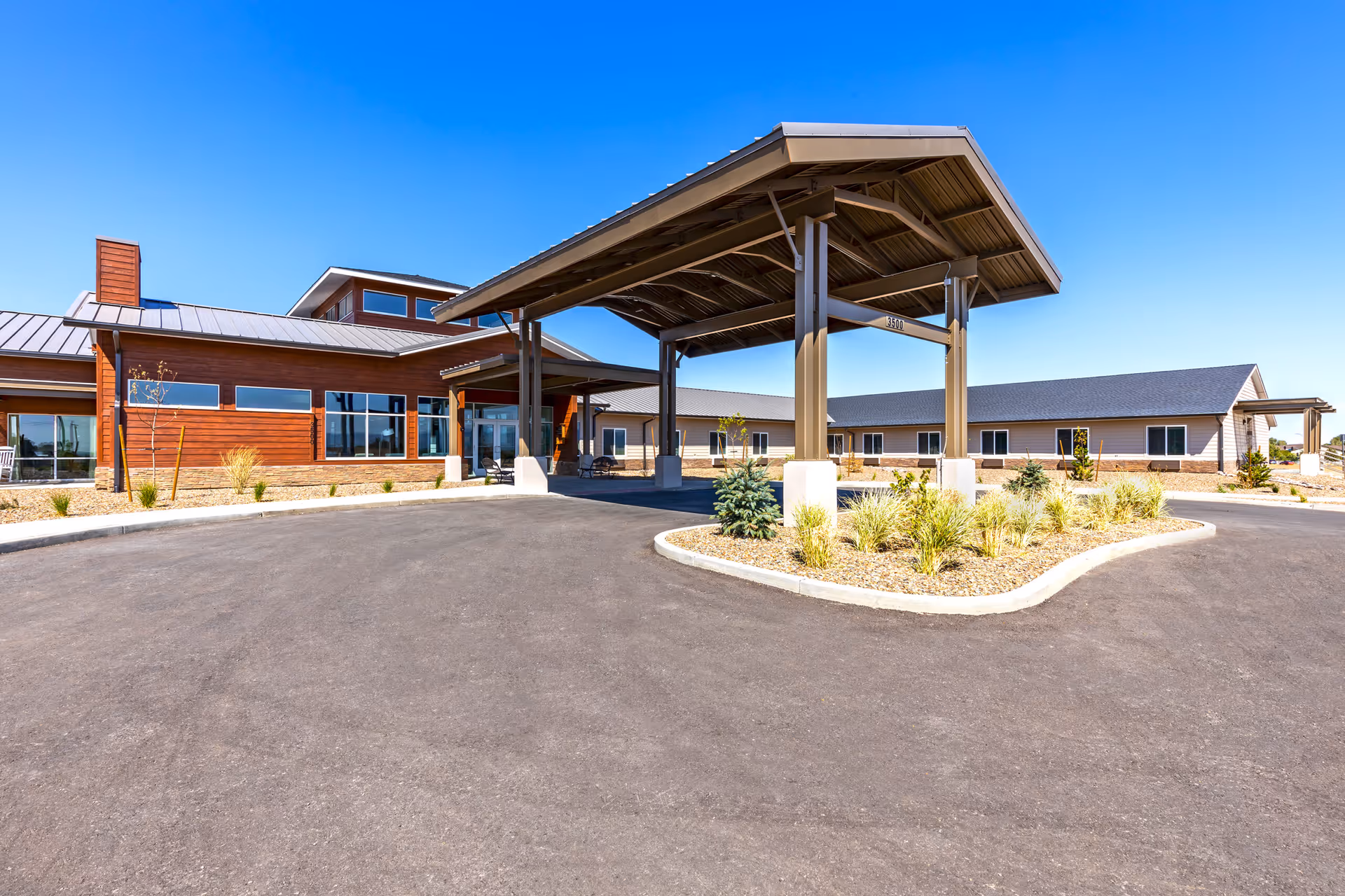Exterior view of The Landings Of Prescott Valley facility showing a large covered entrance with a driveway, landscaped areas with small plants and shrubs, and a clear blue sky.