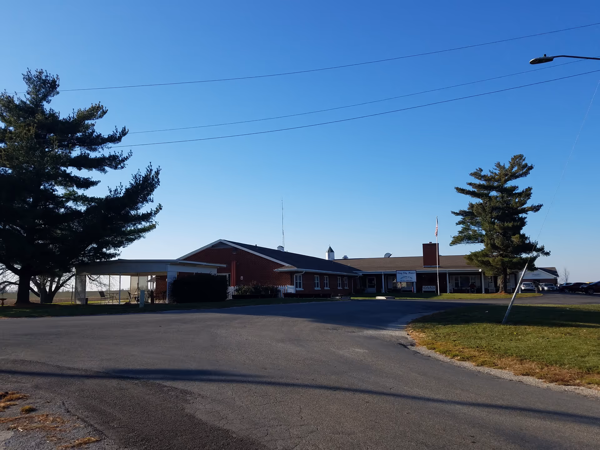 A single-story brick senior living facility sits along a paved road with trees and clear sky.