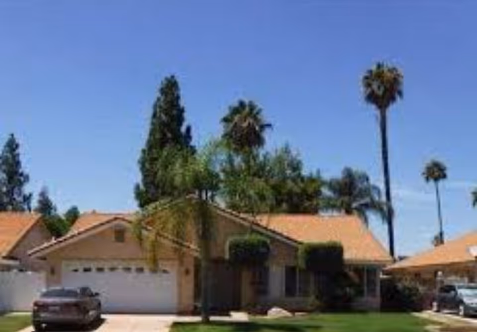 Single-story beige residential building with a tiled roof, driveway, and palm trees under blue sky.