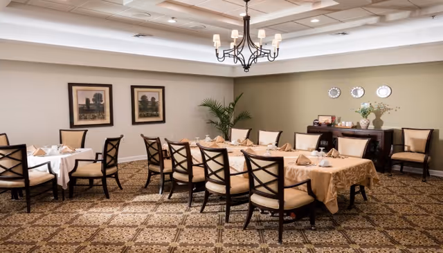A formal dining room with two tables covered in beige and white tablecloths, set with napkins, cups, and plates. The room features cushioned chairs with dark wooden frames, a patterned carpet, framed artwork on the walls, a chandelier hanging from the ceiling, and a sideboard with decorative plates and a flower arrangement.