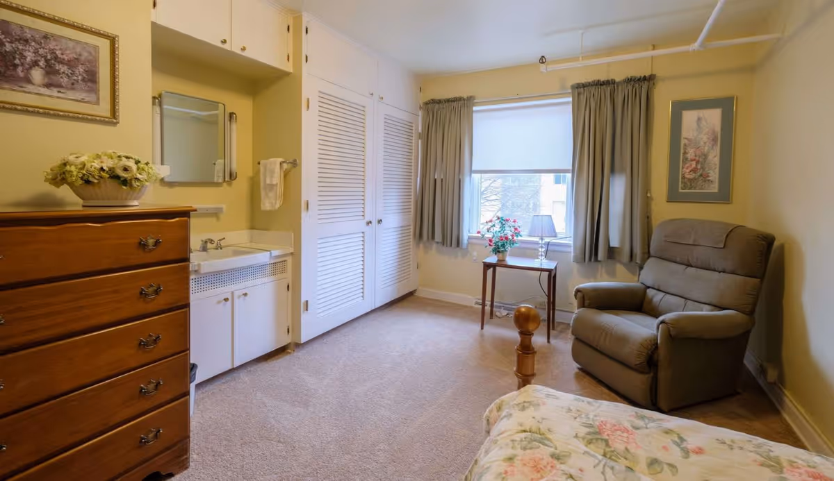 A cozy bedroom in a senior living facility with a floral bedspread, a wooden dresser with a flower arrangement on top, a small sink with a mirror above it, a window with curtains, a small table with a lamp and flowers, and a comfortable armchair.
