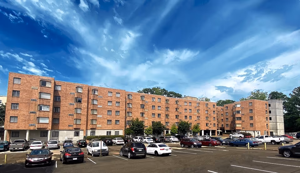 Brick apartment complex with multiple windows and a spacious parking lot under blue sky.