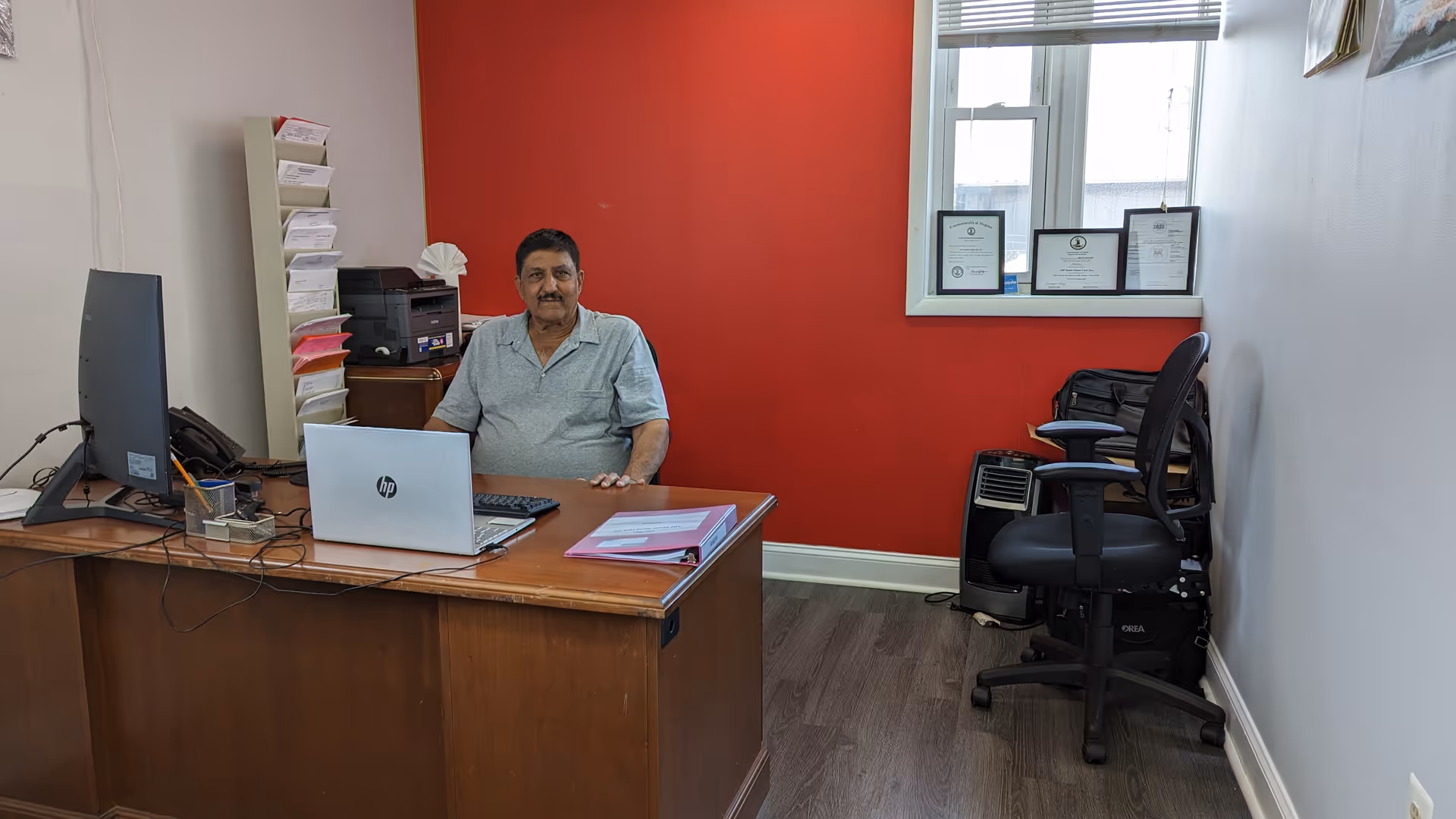 A man sits at a wooden desk in a red-walled office with a laptop.