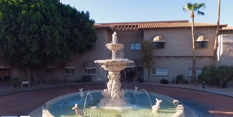 A tiered stone fountain spraying water in a circular pool in a courtyard before a two-story stucco building with palm trees.