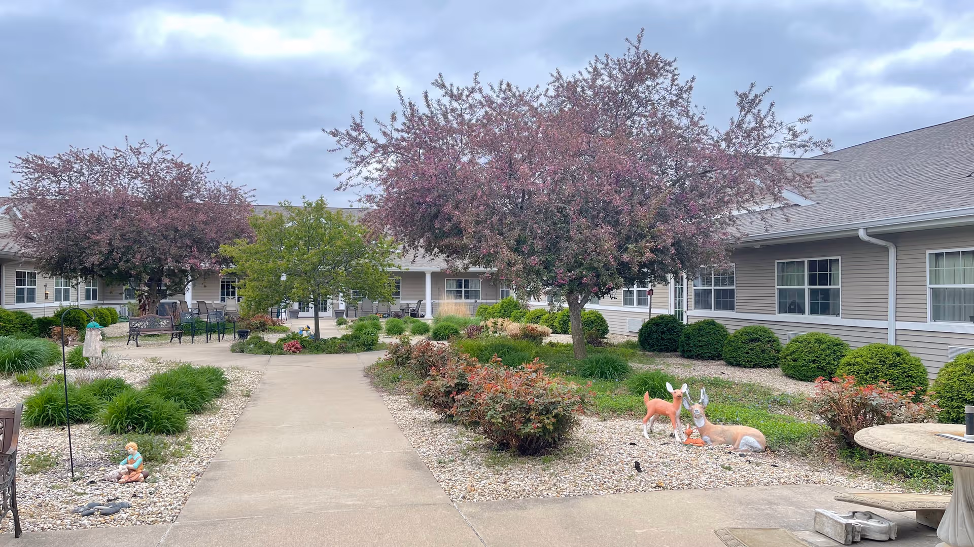 WoodRidge Supportive Living Geneseo courtyard with paths, trees, shrubs, benches, and decorative statues.