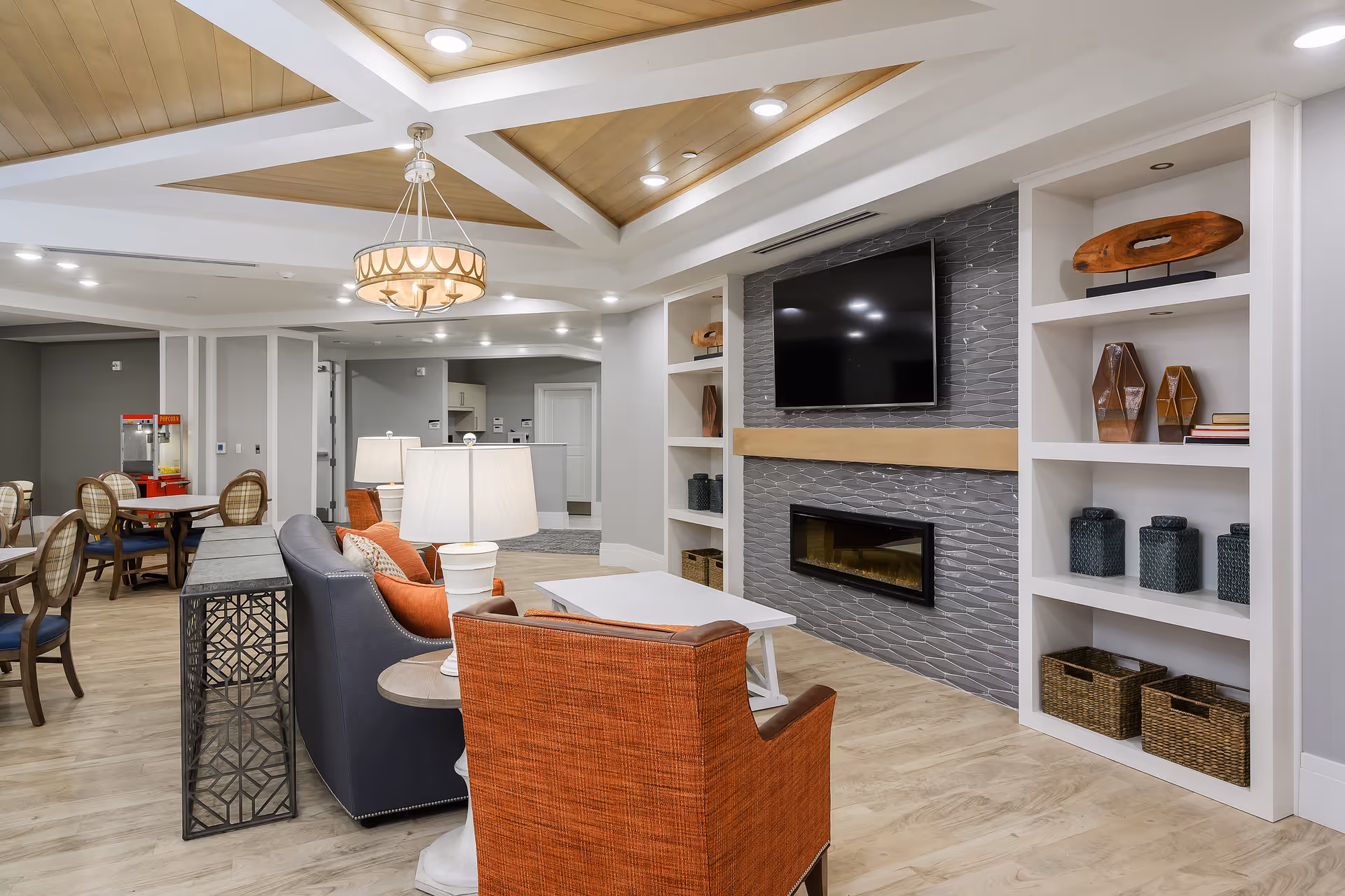 A modern and cozy living room area in a senior living facility featuring a gray couch with orange pillows, an orange armchair, a white coffee table, and a side table with a lamp. The room has built-in white shelves with decorative items, a wall-mounted flat-screen TV above a sleek electric fireplace with gray tile surround, and a ceiling with recessed lighting and a decorative chandelier. In the background, there is a dining area with tables and chairs and a popcorn machine.