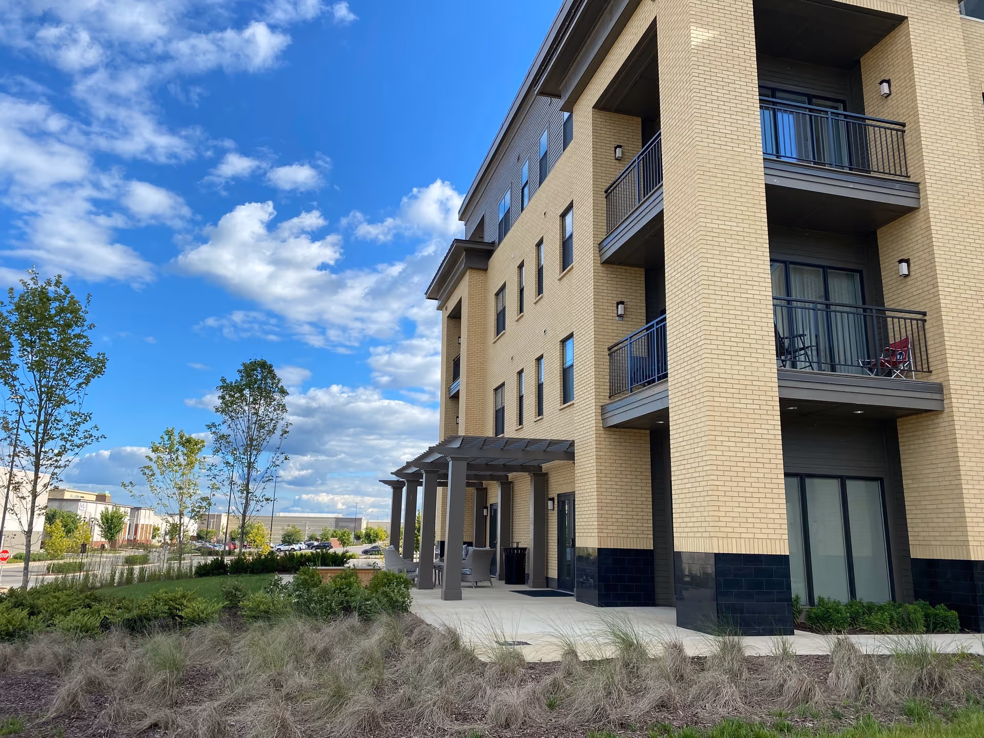 Exterior view of a multi-story senior living facility building with beige brick walls and balconies. The building is surrounded by landscaped greenery and trees under a partly cloudy blue sky.