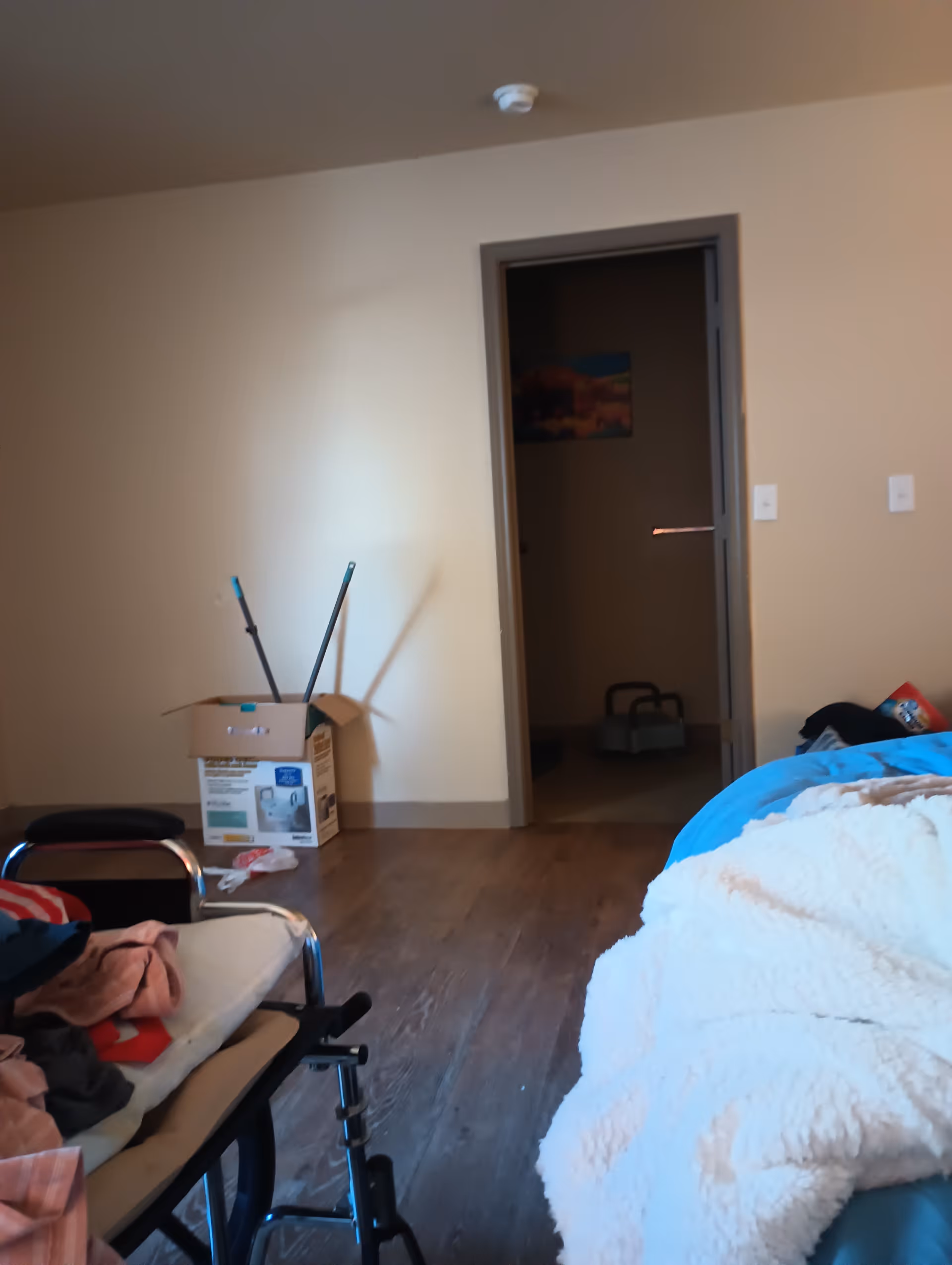 A cluttered patient room with a bed on the right and cleaning supplies near doorway.