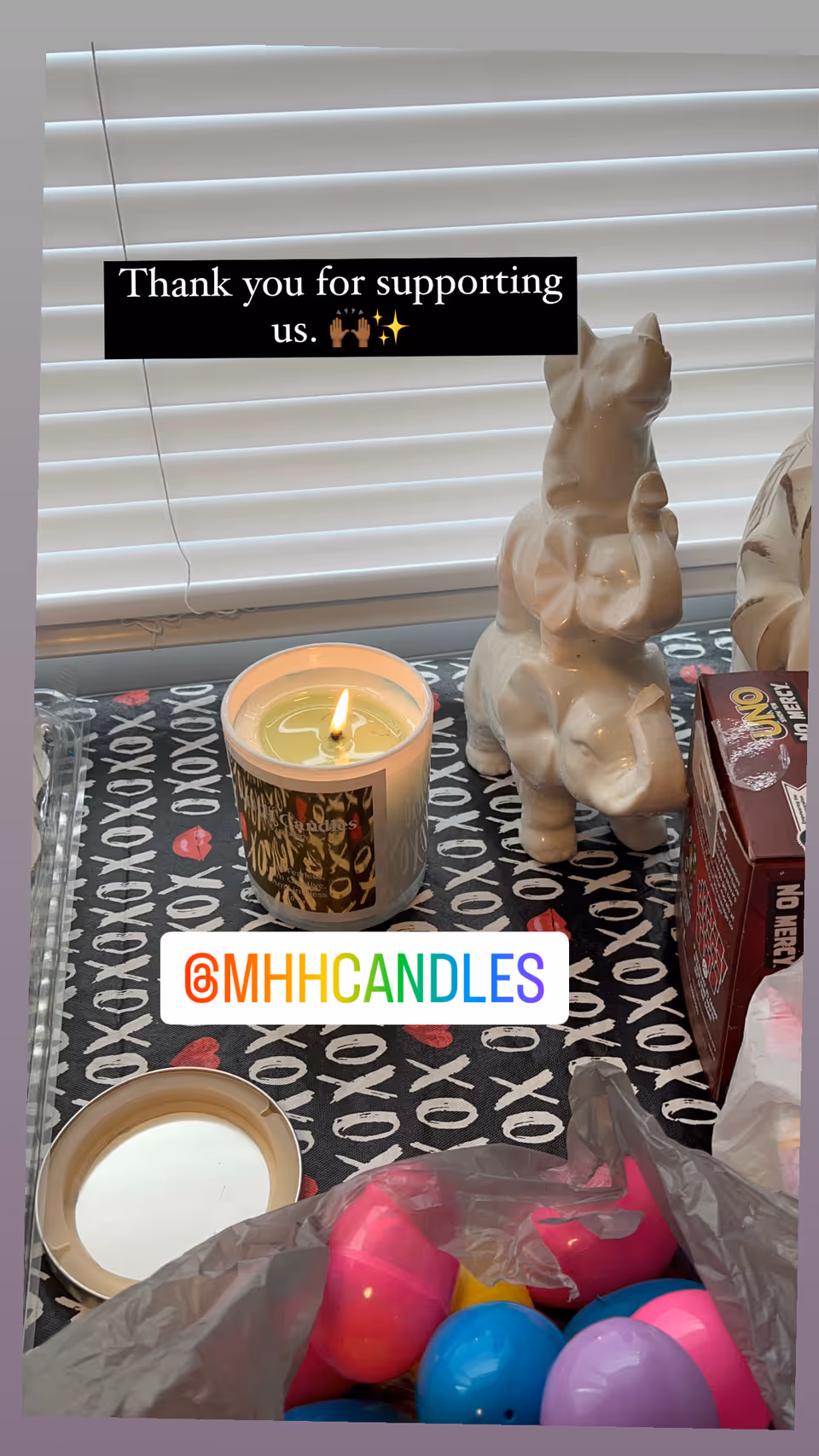 Tabletop with lit candle, ceramic animal figurine, colorful plastic eggs, patterned tablecloth indoors.