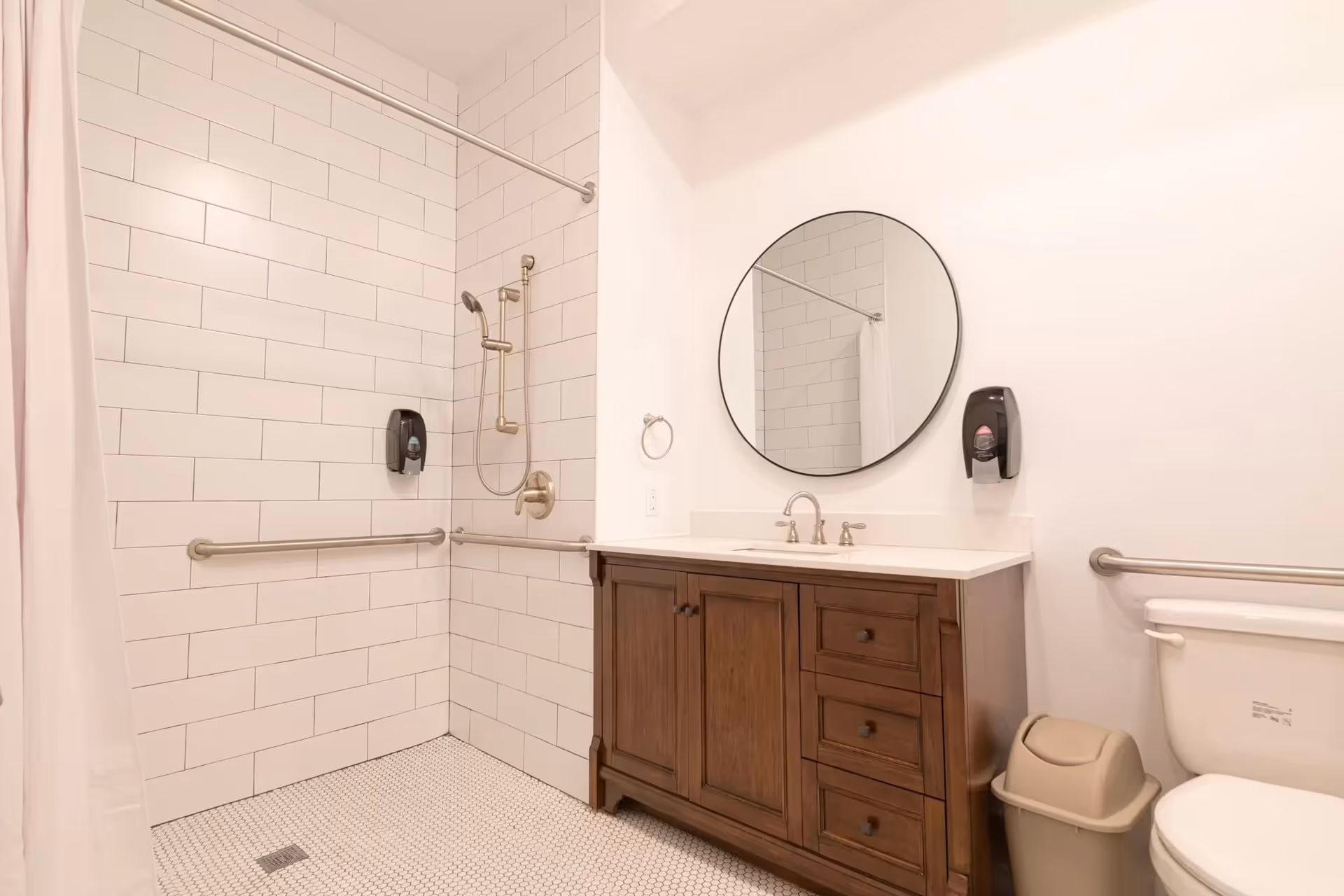 Accessible bathroom with tiled shower, grab bars, vanity, round mirror, and toilet.
