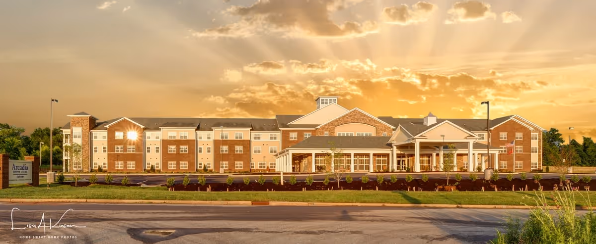 Wide exterior view of a large, three-story senior living facility building at sunset with a well-maintained lawn and parking area in front. The building features a combination of brick and light-colored siding with multiple windows and a covered entrance. A sign on the left reads 'Arcadia Senior Living'.