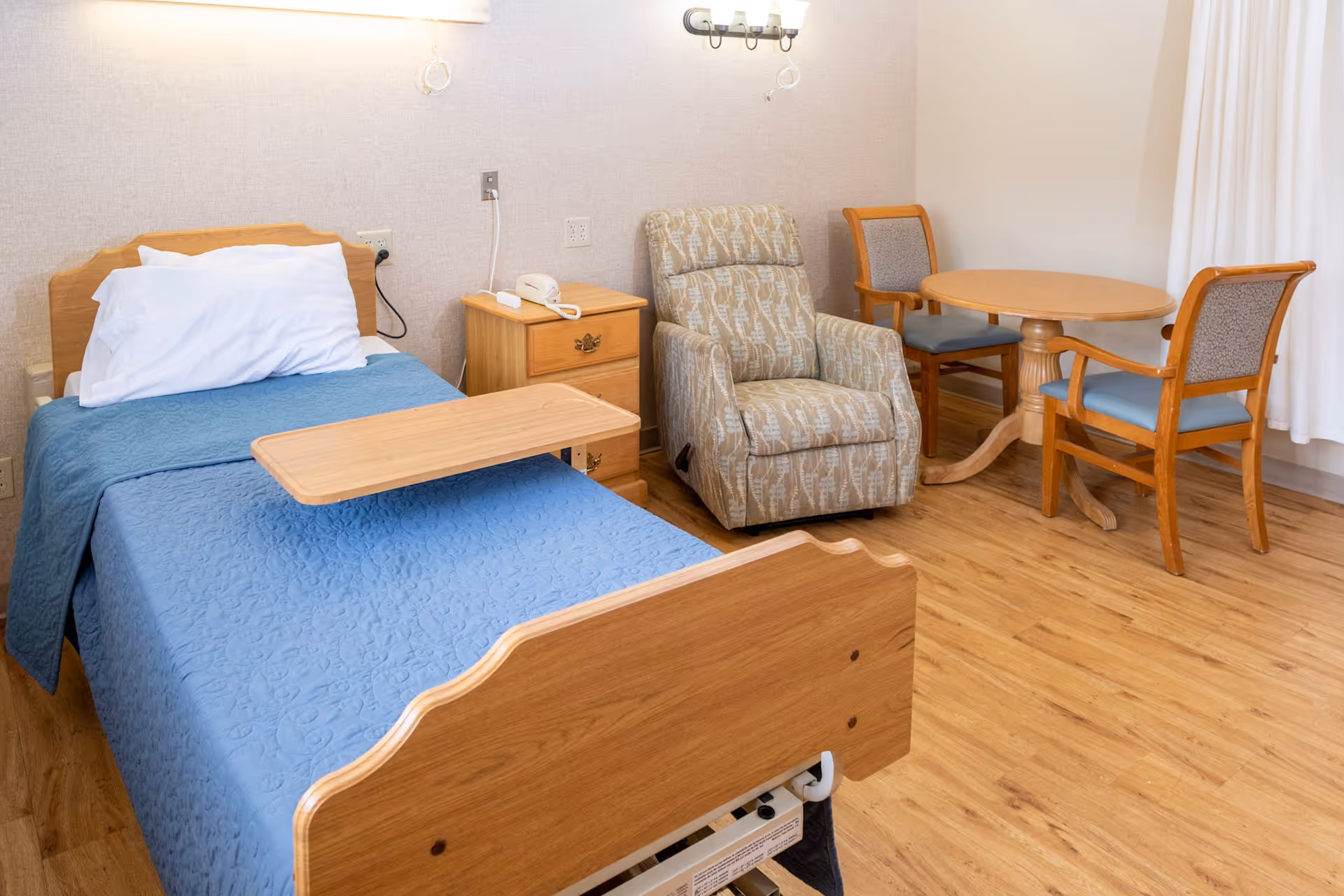 A patient room with a wooden bed, recliner, bedside table, and small dining table.