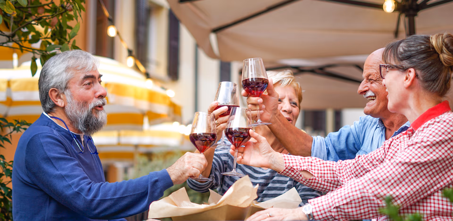 Four older adults sitting outdoors at a table, raising glasses of red wine in a toast, smiling and enjoying each other's company under a canopy with yellow and white striped umbrellas in the background.