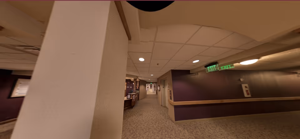 Carpeted interior corridor of a retirement community with purple walls, a large pillar in the foreground, ceiling tiles, and an illuminated EXIT sign.