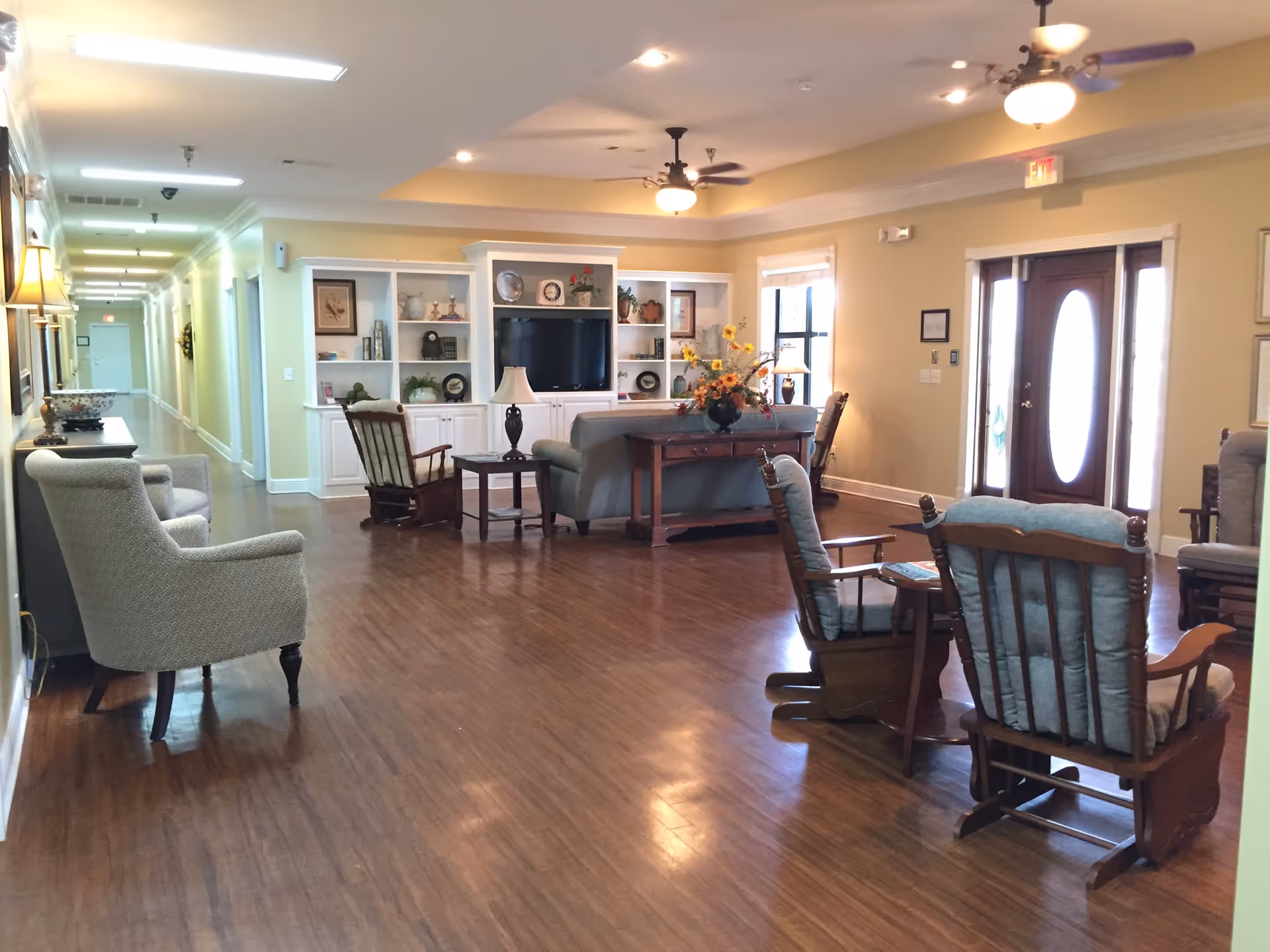 A spacious and well-lit living room area with wooden flooring, several cushioned armchairs and rocking chairs arranged around a central coffee table with a flower vase. There is a large built-in white shelving unit with a television and decorative items against the far wall. The room has ceiling fans with lights, large windows, and a wooden front door with glass panels.