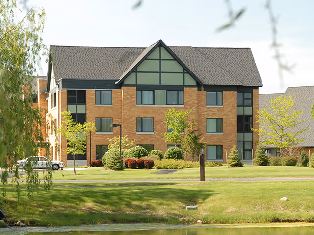 Exterior view of a three-story brick building with green trim and multiple windows, surrounded by green grass, small trees, and shrubs, with a pond in the foreground.