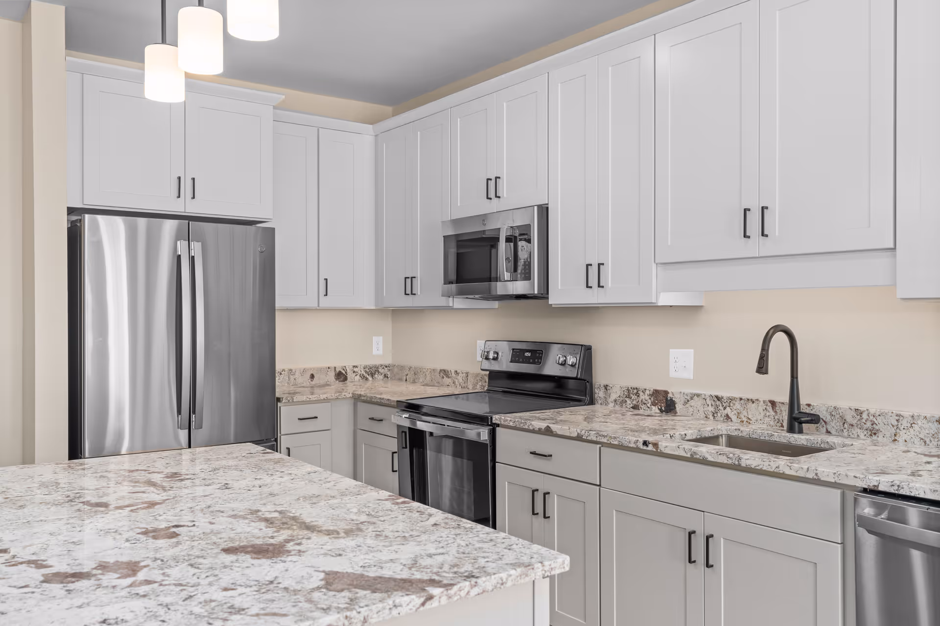Modern kitchen with white cabinetry, granite countertops, stainless steel refrigerator, stove, microwave, dishwasher, and a sink with a black faucet. Three pendant lights hang from the ceiling above the counter.