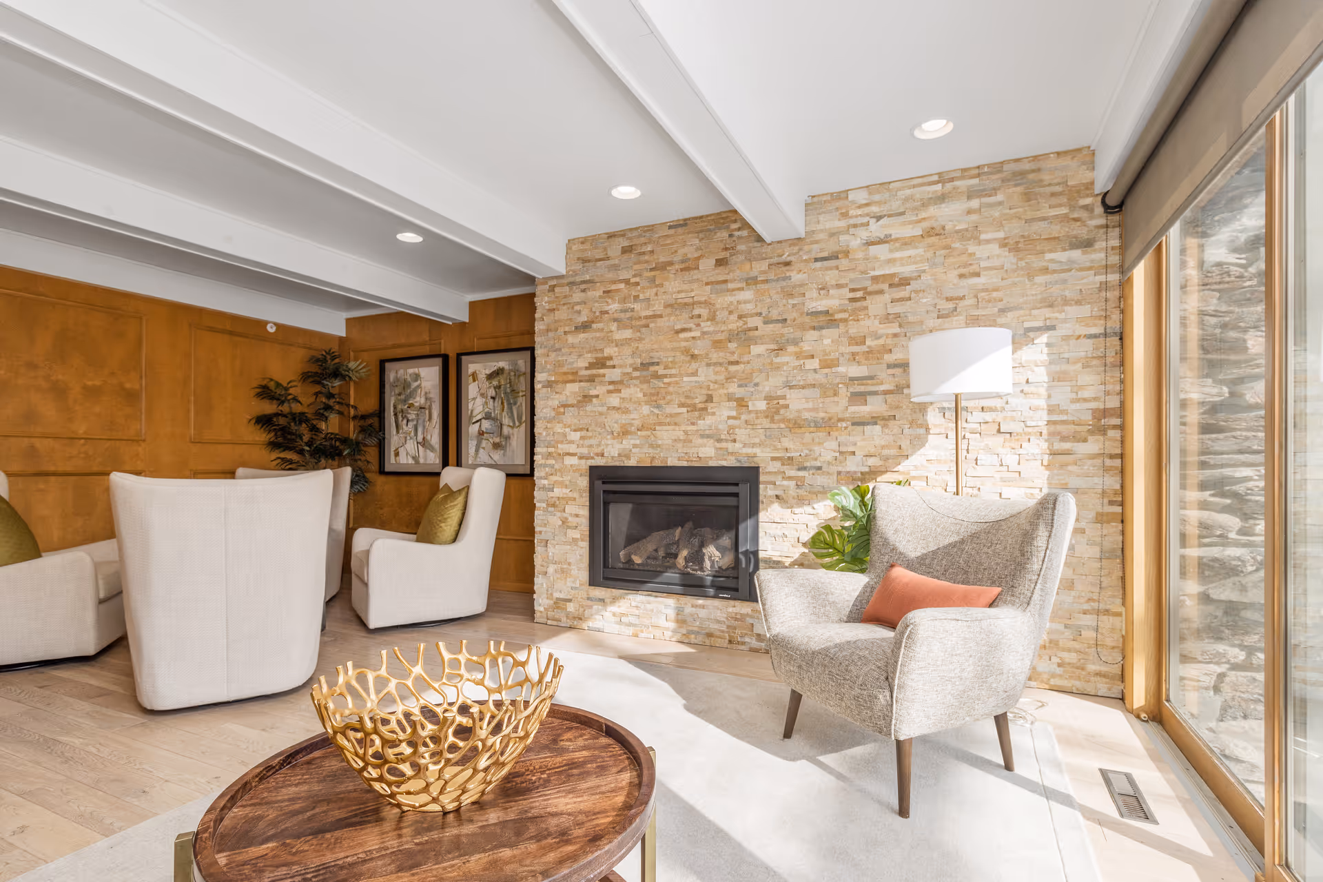 A bright and cozy living room with a stone accent wall featuring a built-in fireplace. The room has light-colored wooden floors, a beige armchair with a coral pillow, a floor lamp, and a wooden coffee table with a decorative golden bowl. There are also two white armchairs with green pillows, framed artwork on the wood-paneled wall, and large windows letting in natural light.