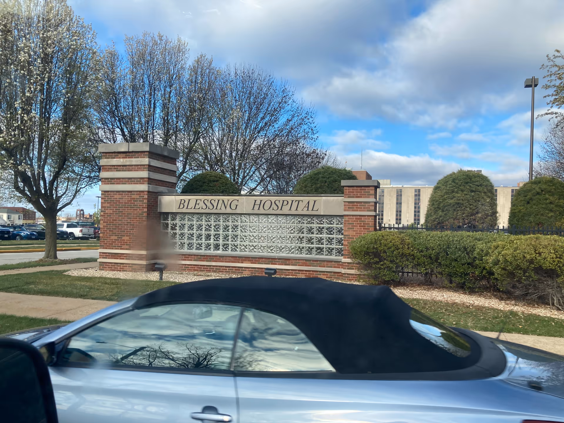 Brick hospital entrance sign reads Blessing Hospital with trees, hedges, and a parked convertible.