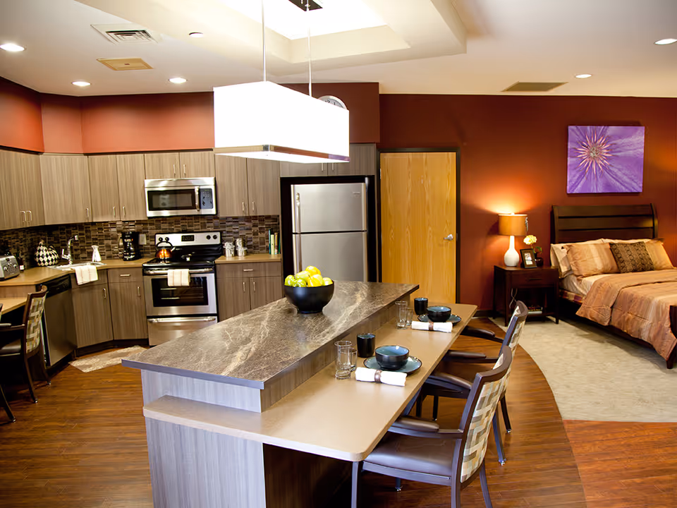 A modern studio apartment interior with a kitchen and bedroom area. The kitchen features wooden cabinets, stainless steel appliances including a refrigerator, stove, microwave, and dishwasher. A large island with a marble countertop has three place settings with bowls, glasses, and napkins. The bedroom area has a bed with beige and brown bedding, a nightstand with a lamp and a small flower vase, and a purple flower artwork on the wall. The walls are painted a warm reddish-brown color and the floor is a combination of wood and carpet.