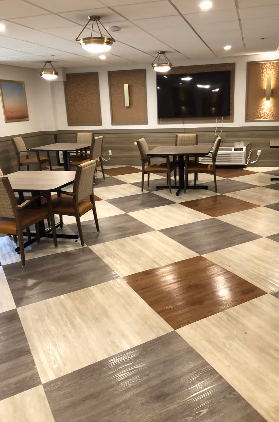 A dining area with several square tables and chairs arranged on a checkered floor with brown, beige, and gray tiles. The walls have decorative panels and a large flat-screen TV mounted on one side. Ceiling lights hang above the tables, and there is an air conditioning unit below the TV.