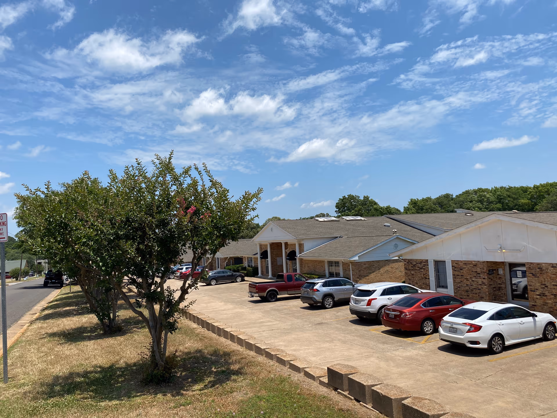 Front exterior of the Avir at Rose Trail senior living building with a row of parked cars, trees, and a blue sky.